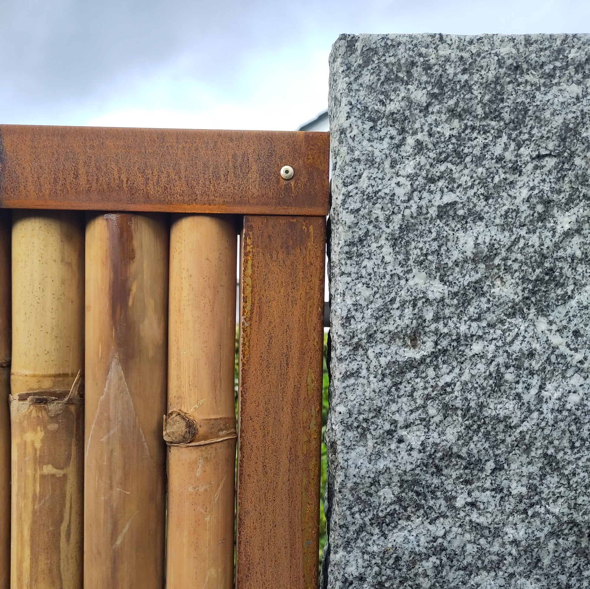 Obere Ecke eines montierten Bambus Zaunfeldes mit umlaufendem Rahmen aus Cortenstahl - das seitliche Rahmenprofil wurde an einer Granit-Palisade befestigt